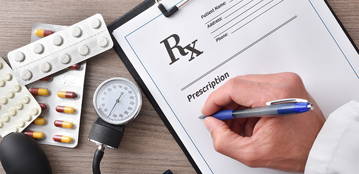 A hand holding a pen writing on a piece of paper on a clipboard; medicine tablets and a blood pressure monitor