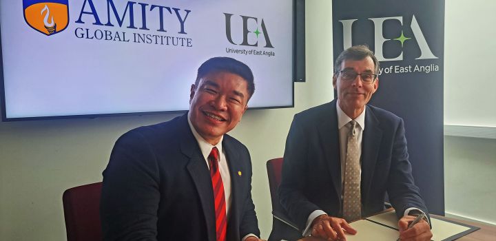 Prof Leon Choong and Prof Stephen McGuire, both smartly dressed in suits and ties, sign the contract documents in front of the logos of Amity and UEA.