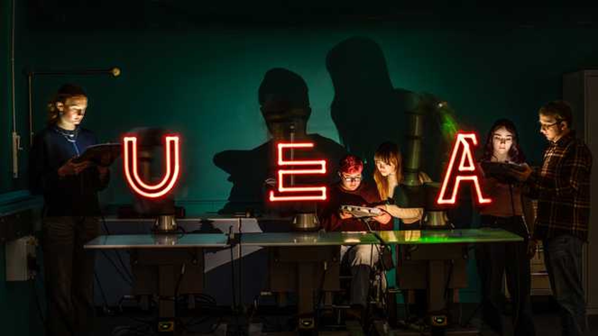 People in a dark room illuminated by neon lights that spell out U E A
