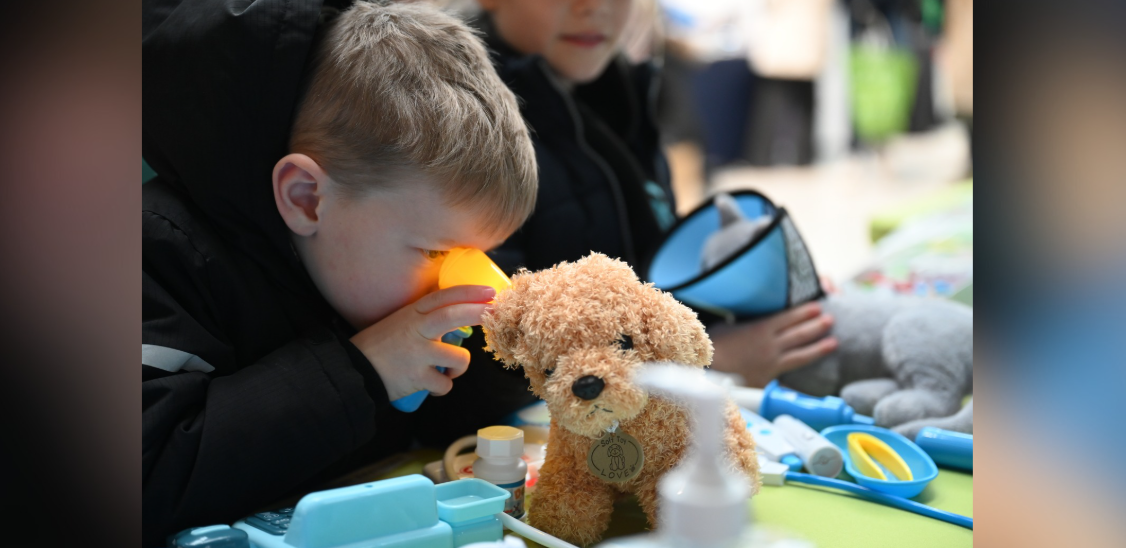 A boy looking closely at an orange, stuffed dog teddy through an orange device, while someone in the background is holding a grey cat teddy