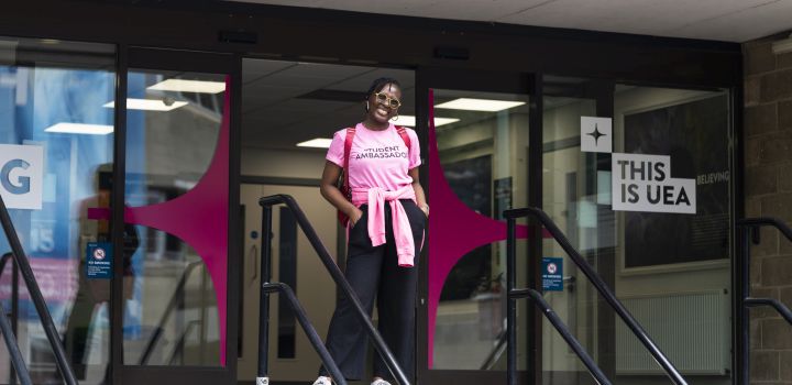 A student ambassador in a pink branded T-shirt stands in front of some open sliding doors, with a sign on the right saying "This is UEA"