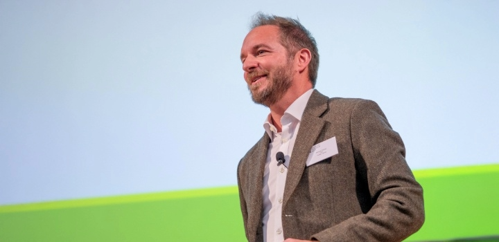 A man wearing a jacket and shirt stands in profile against a white and green background