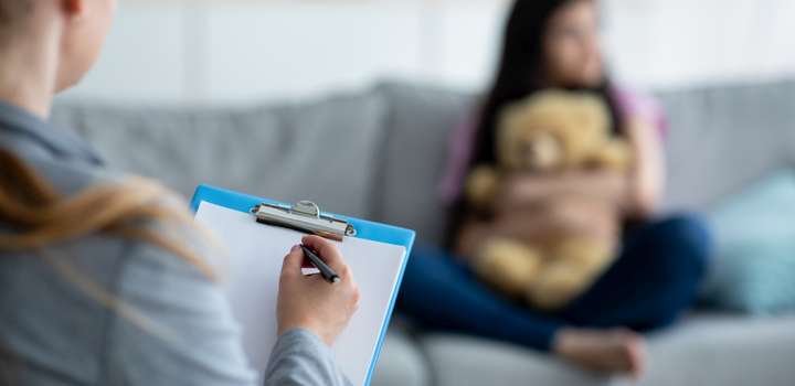 A young girl sits on a sofa cuddling a teddy bear while talking to a mental health professional