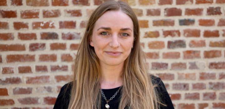 Martha Loader smiling at the camera. She is wearing a black top and standing in front of a red brick wall.