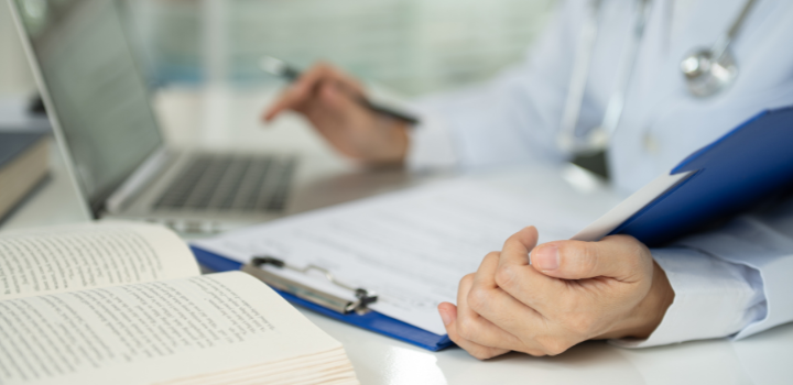 A medical professional looking at a clipboard and laptop on a desk