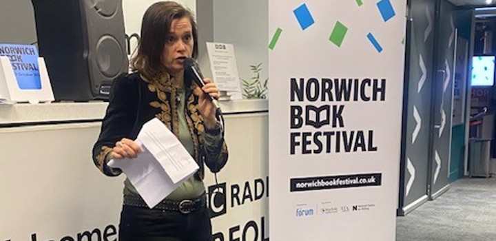 Prof Jean McNeil, Director of Creative Writing, at UEA,  speaking on  a microphone in front of a Norwich book festival banner
