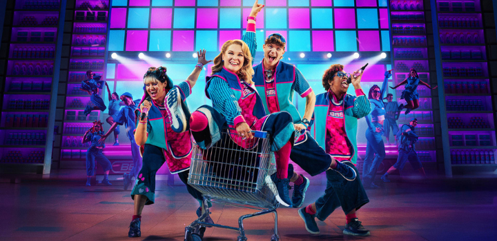 A photo of the cast of the musical Here & Now, a person is sat in a trolley being pushed, while two others hold microphones next to them