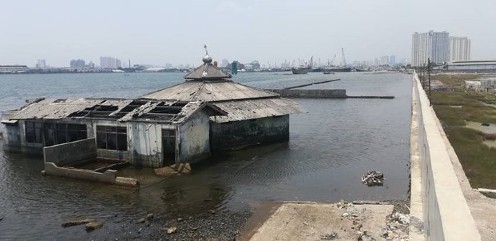 Remains of a building surrounded by sea water next to a sea wall, with a city in the background