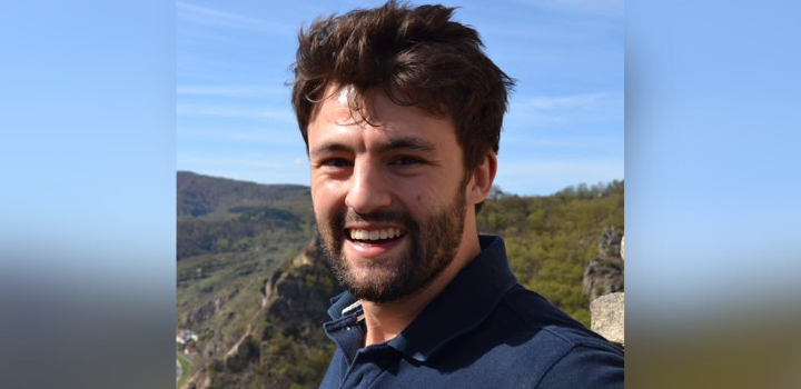 A man smiling at the camera, there's a blue sky and green hills behind him