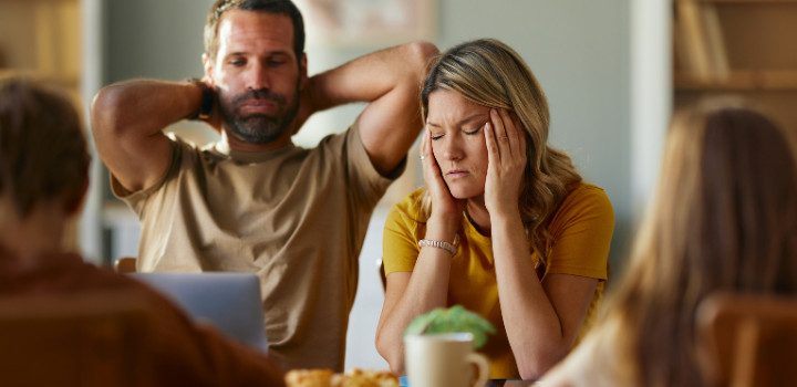 A stressed man and a stressed woman sat at a table, two blurry figures of children sat in front of them