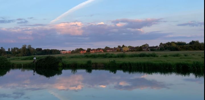 The Fens, a serene body of water reflects the flat and grassy landscape and purple-hued sky.