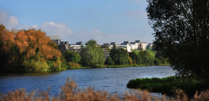 View across the lake at UEA, with trees, lakeside habitats and campus buildings visible beyond the water.