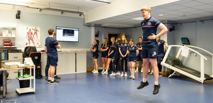 A student jumps in the air with mini sensors attached to various parts of his body, while his peers look at a television screen where his movement is being mapped.