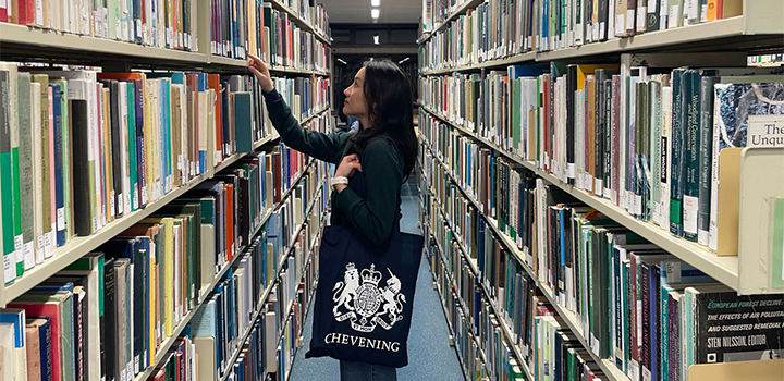 Kulasinee Pipattanasookmongkol standing in the middle of two bookshelf aisles at the UEA Library, reaching for a book to her left and holding a Chevening bag