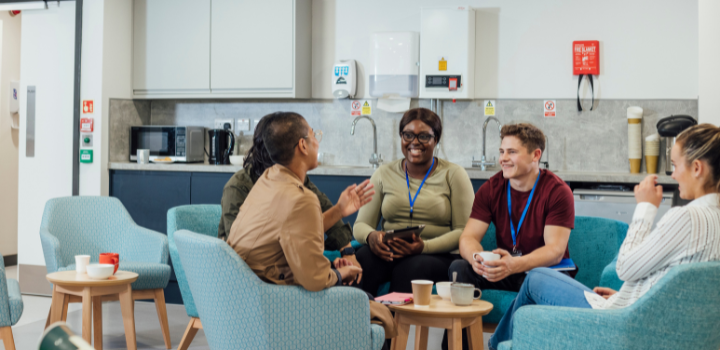 A group of healthcare professionals sitting in chairs around a wooden table in a staff common room