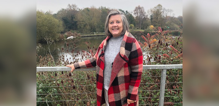 Prof Sally Hardy smiling and wearing a red checked coat, as she stands in front of a lake