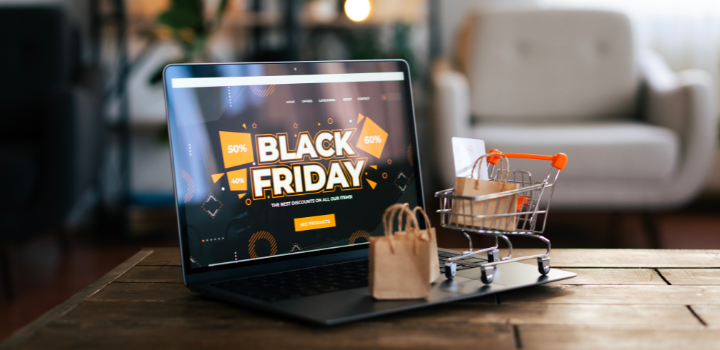 A laptop on a table, it has a website on it displaying 'Black Friday' with a miniature shopping trolley and some cardboard shopping bags