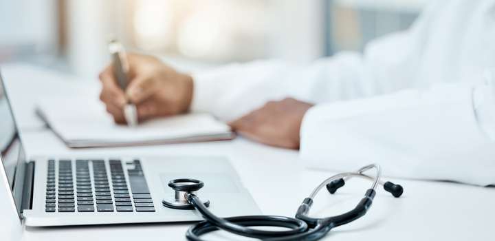 A researcher in a white lab coat makes notes, while in the foreground is a laptop computer and a stethoscope.