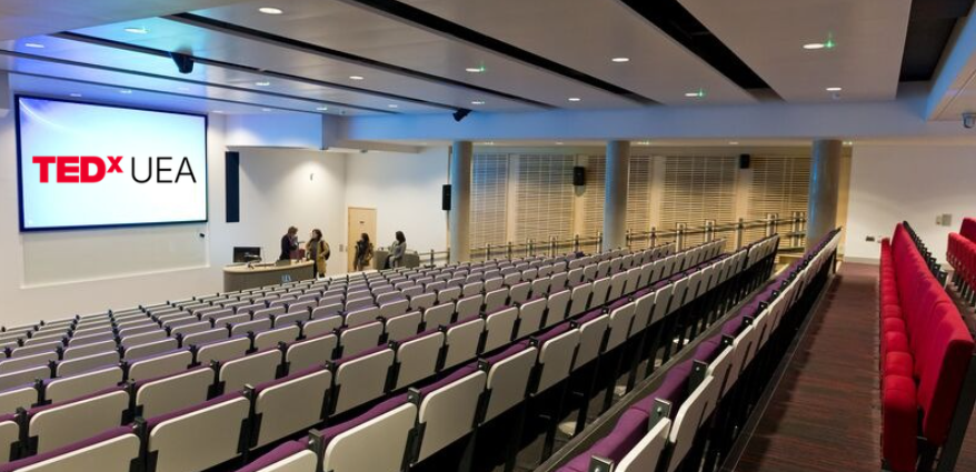 An empty lecture theatre with four people speaking amongst themselves at the front, a red and black TEDxUEA logo is on a whiteboard beside them