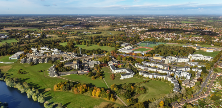 An aerial view of the UEA campus, with green trees and grounds and a blue sky in the background