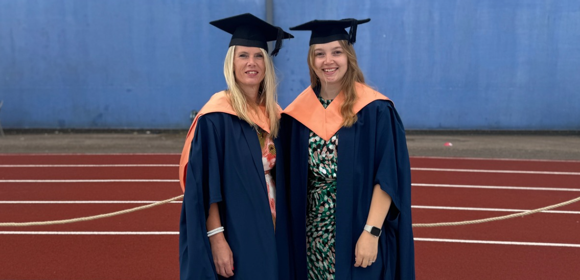 Two graduates in navy robes and coral collars smiling at the camera, they are wearing graduation caps and standing in front of a blue wall