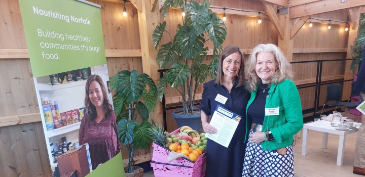 Anna Sweeting and Dr Sarah Hanson stand next to a basket of fruit and vegetables and a banner which says Nourishing Norfolk