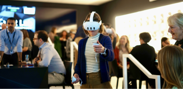 A woman standing up with a VR headset on, there's a small crowd around her.