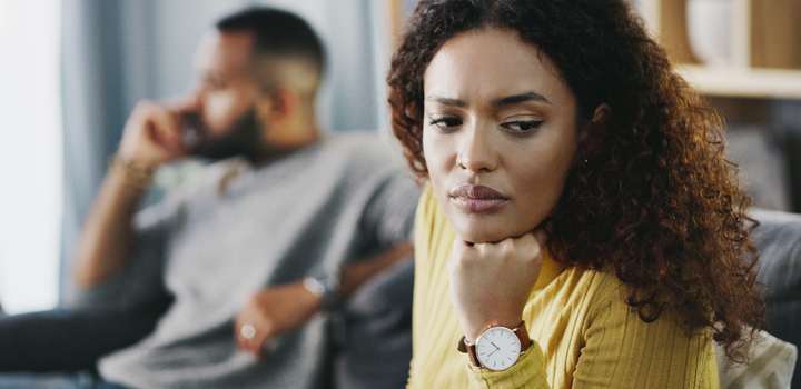 A woman looks pensive and troubled in the foreground as her partner can be seen sitting on the same sofa in the background.