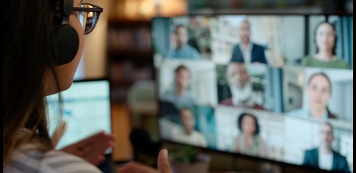 A woman, wearing headphones, sits in front of a computer where the screen is a of a group video call with 9 different people on it.