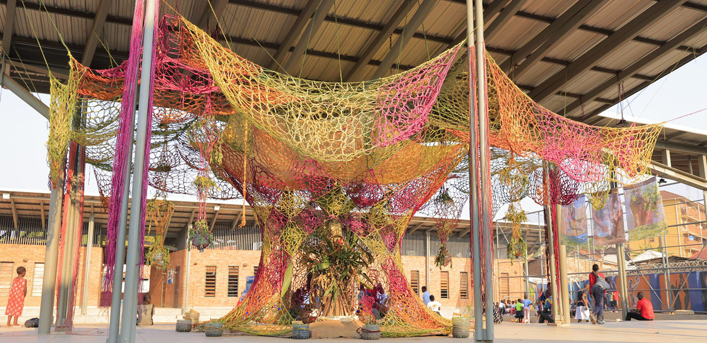 A large, colourful net 'tree of life' art installation hangs in a courtyard as people interact with it.