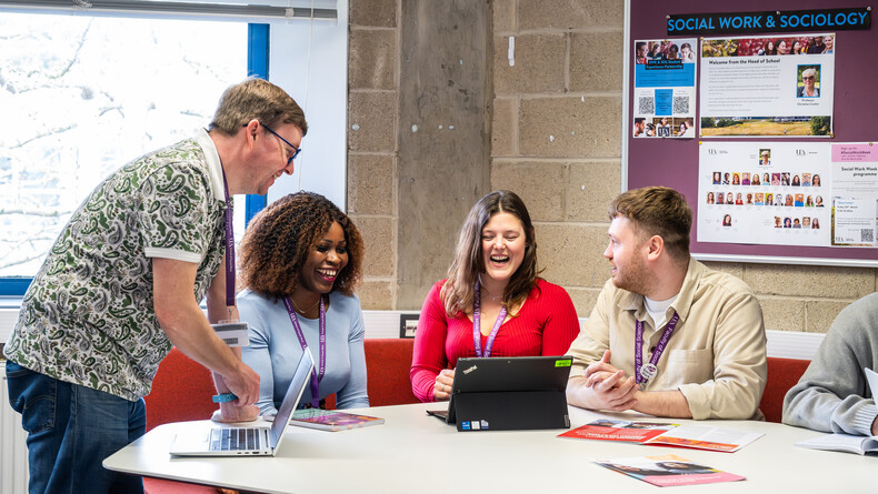 Three students are in a Social Work and Sociology social space, chatting with each other while talking to a lecturer, all wearing purple lanyards.