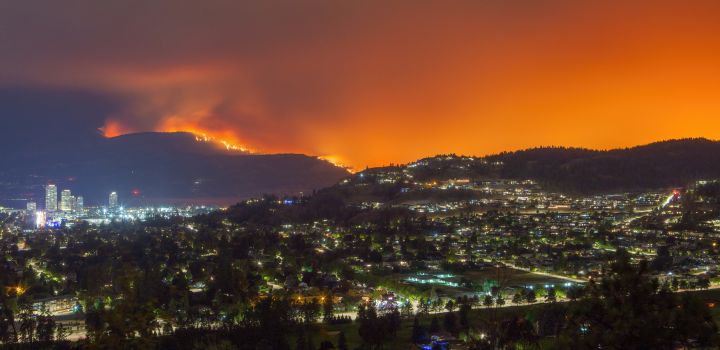 A wildfire burns on a mountainside near Kelowna illuminating the night sky with an orange glow while city lights shine below.