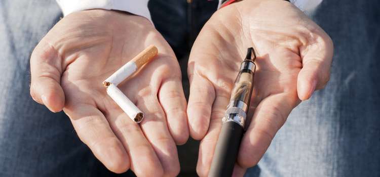 A close up of a person's hands holding a broken cigarette in the left one and a vape in the right one, symbolising the choice between the two.