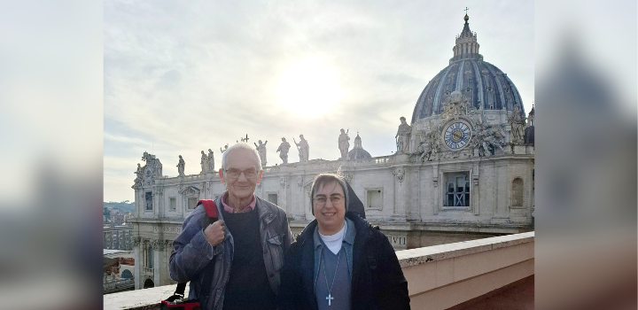 Prof Robert Sugden and Sister Alessandra Smerilli standing on a roof terrace in Vatican City, with the dome of St Peter's Basilica visible behind them.