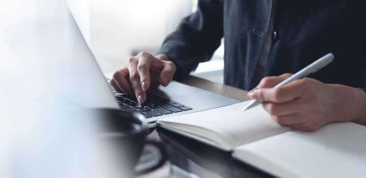 A person seated at a desk, one of their hands is using a laptop while the other is writing with a pen in a notebook