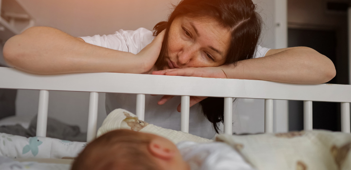 Young mother feeling exhausted and overwhelmed. Resting her head on the edge of a baby's crib while looking down at the sleeping infant