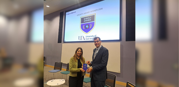 A woman hands a small award to a tall man in front of a big screen displaying the name of the award.