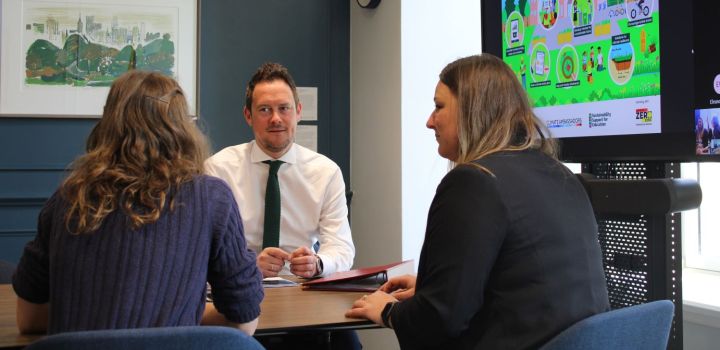 The UK Minister for Early Years, Stephen Morgan MP,  sits facing the camera at a desk, wearing a white shirt with a dark tie, while talking to two seated women