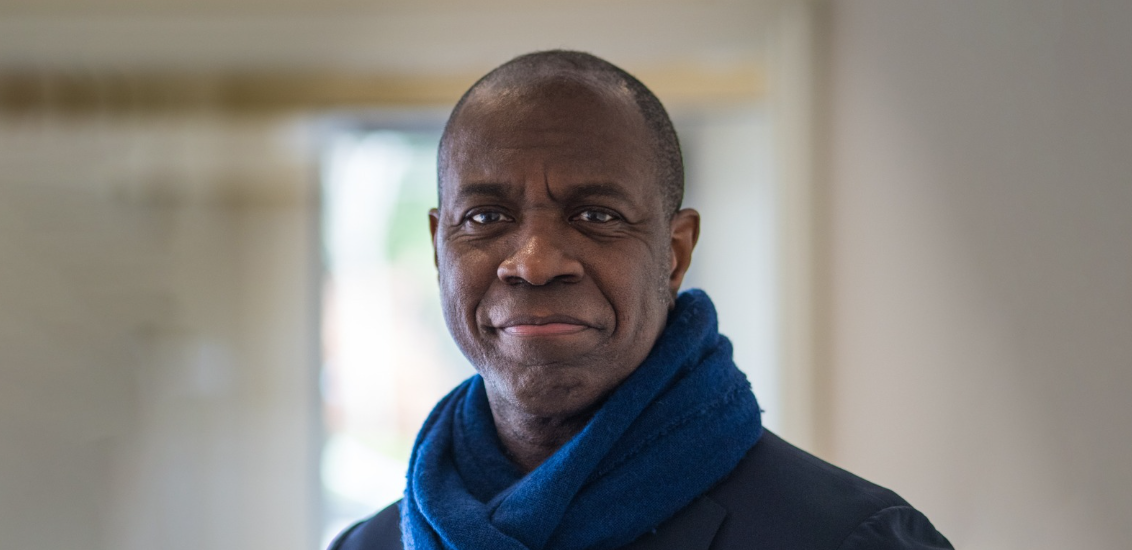 Clive Myrie smiling at the camera, wearing a blue scarf and black jacket, in front of a brightly-lit window in the background