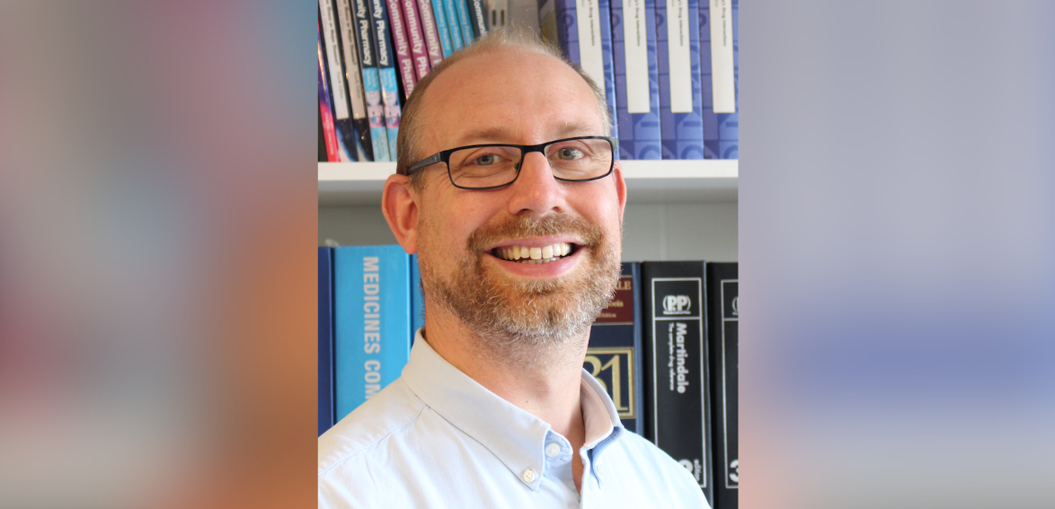 A headshot of a man in glasses smiling at the camera, standing in front of a bookcase filled with textbooks
