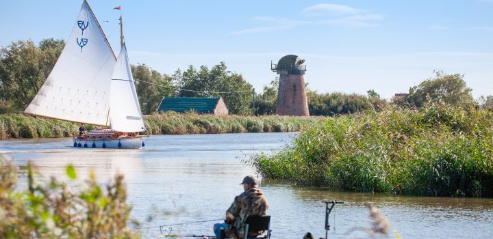 An angler sits with his fishing rod at Upton Dyke on the Broads. In the background is a former windmill, which no longer has its sails, and a sailing boat, which is on the water.