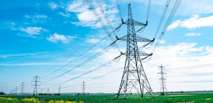 A green field with a large electricity pylon and other pylons in the background, against a blue sky with white clouds