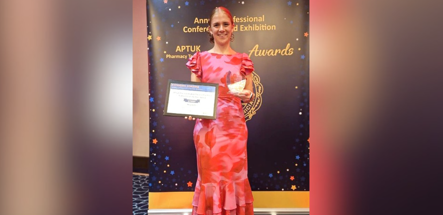 A smiling woman in a red and pink floral long dress, holding an award certificate in front of a black awards banner