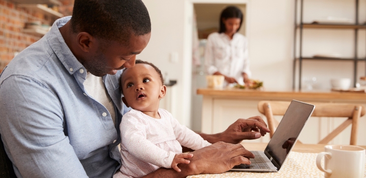 Father and baby daughter use laptop as mother prepares meal