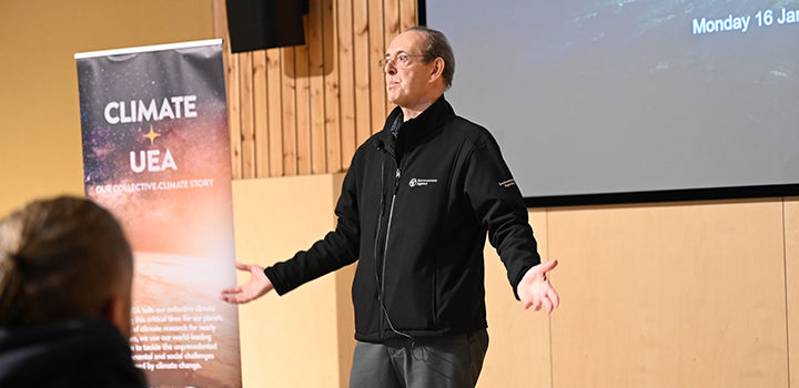 James Bevan presenting at UEA. He is wearing glasses, a black jacket and grey trousers.