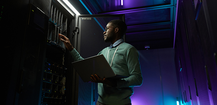 A man inspects a computer server while holding a laptop.